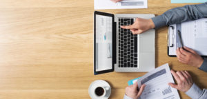 Aerial view of table with white laptop on it. 2 people working together with only view of their hands and some paper work. Person pointing to the screen on laptop